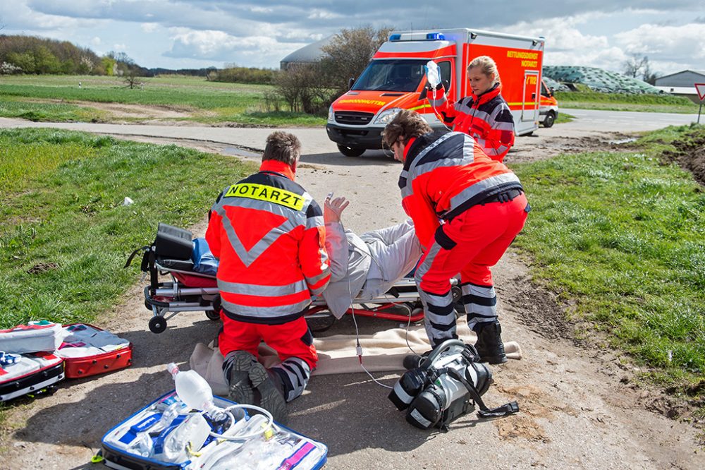 Straßenkreuzung in einer ländliche Gegend. Einsatzfahrzeuge und Rettekräfte im Einsatz an einer Krankentrage.