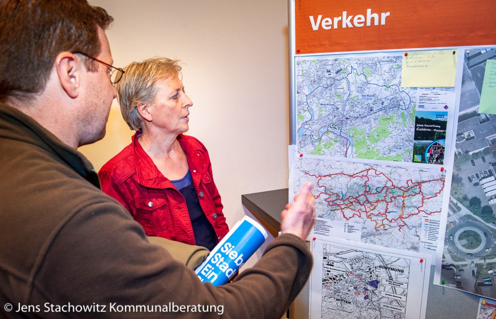 Mitarbeiterin der Stadtverwaltung und Bürger vor einer Plakatwand